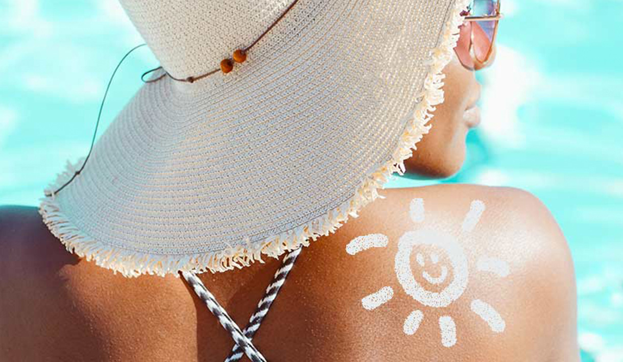 Eine Frau am Meer mit Hut und einer Sonne aus Sonnenmilch auf dem Rücken.